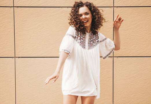 Beautiful smiling model with afro curls hairstyle dressed in summer hipster white dress.Sexy carefree posing in street near yellow wall.Positive woman having fun