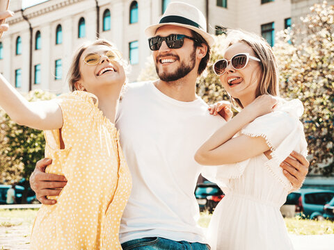 Group of young three stylish friends in the street.Man and two cute female dressed in casual summer clothes.Smiling models having fun in sunglasses.Women and guy making photo selfie on smartphone