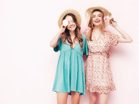 Two young beautiful smiling brunette hipster female in trendy summer dresses. Sexy carefree women posing near pink wall. Positive models Holding flowers. Cheerful and happy. In hats