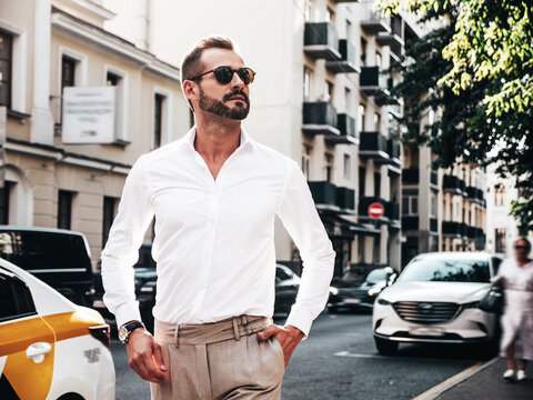 Portrait of handsome confident stylish hipster lambersexual model.Modern man dressed in white shirt and trousers. Fashion male posing in the street background in Europe city at sunset. In sunglasses