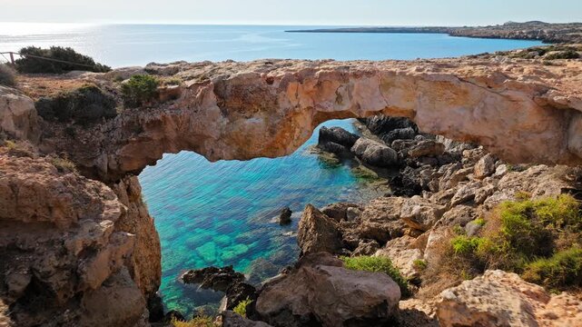 Aerial view looking through the Crow&rsquo;s Arch rock opening at Cape Cavo Greco, Cyprus. The natural stone frame reveals clear blue sea and rugged coastal formations.