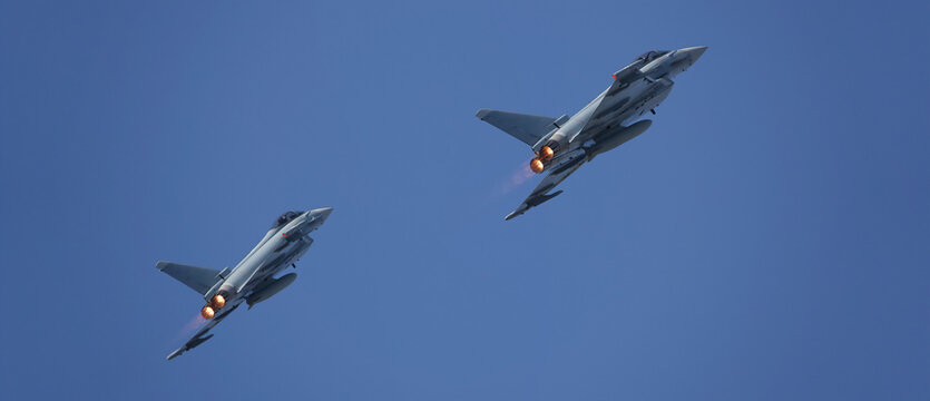 Two Eurofighter typhoon multirole combat aircraft flying in close formation against a clear blue sky, their afterburners glowing brightly.