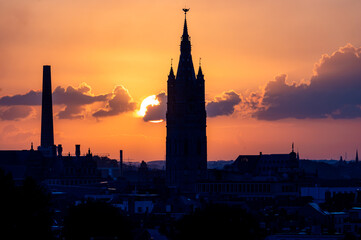 A Gothic church tower of Ghent stands in silhouette in the warm morning glow with a factory chimney nearby in Belgium. © Oksana