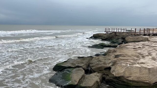 Foamy waves keep washing across rugged rocky shoreline beside small wooden viewing deck, with wet stone ledges, green algae patches, calm open sea, and pale overcast sky shaping quiet cool coastal