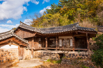 Obraz premium Scenic Exterior View of Traditional Korean Hanok Architecture Featuring Tiled Roofs, Earthen Wall, and Vibrant Autumn Foliage Under Blue Sky