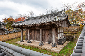 Obraz premium Exterior View of Traditional Korean Hanok Building Featuring Wooden Porch, Curved Tiled Roof, and Foreground Garden Plants Under Sky with Clouds