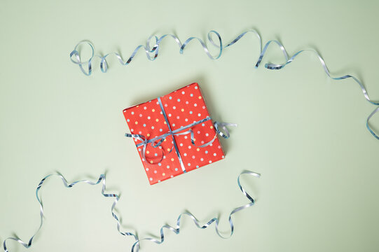 A small gift wrapped in red polka dot paper sits on a mint green background, surrounded by silver curled ribbon.