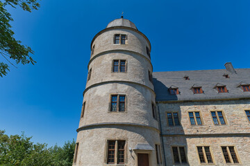 Obraz premium A low-angle shot captures the Wewelsburg Castle. A prominent, multi-story round stone tower with a conical roof stands against a clear blue sky, adjacent to a larger stone building.