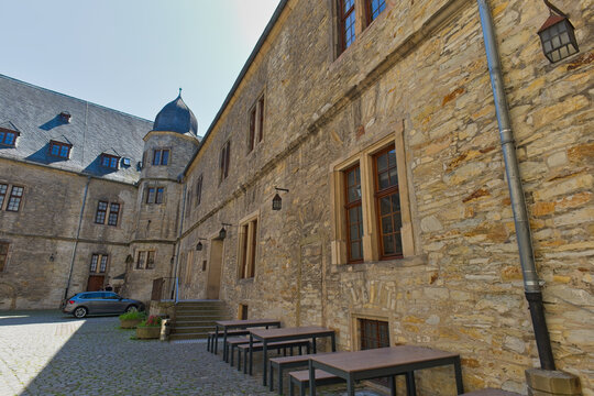 An exterior view of historic Wewelsburg Castle, showing a stone courtyard with outdoor tables and benches. A larger castle building with a dome roof stands in the sunny background.