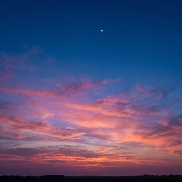 Dramatic sunset sky featuring vibrant pink and orange clouds with a small crescent moon visible in the deep blue twilight atmosphere above a dark silhouette of a distant horizon line