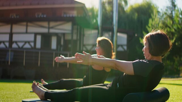 Elderly women practicing slow motion Pilates exercises outdoors on yoga mats, stretching arms and strengthening core muscles together on a sunny day