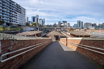 Naklejka premium Red brick pedestrian ramp toward the historic platforms of North Melbourne railway station in Victoria, Australia, with modern high-rise apartments and the Melbourne city skyline in background