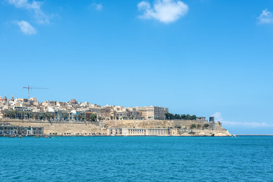 Panoramic view of Valletta, Malta, with historic limestone fortifications, waterfront architecture, and Grand Harbour under a bright blue sky. Scenic Mediterranean coastal city landscape.