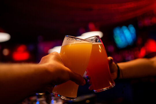 Two people clink glasses of hazy craft beer in a toast at a dimly lit bar with red neon lights.