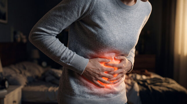 Person holds their stomach in pain while sitting on bed in a dimly lit room during evening hours
