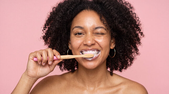 Woman brushes her teeth in front of a pink background while smiling and enjoying the moment in a bathroom