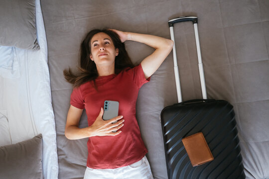 Woman lying on a hotel bed with a smartphone, suitcase, and passport holder, looking up thoughtfully, dreaming about upcoming vacation or business trip adventures