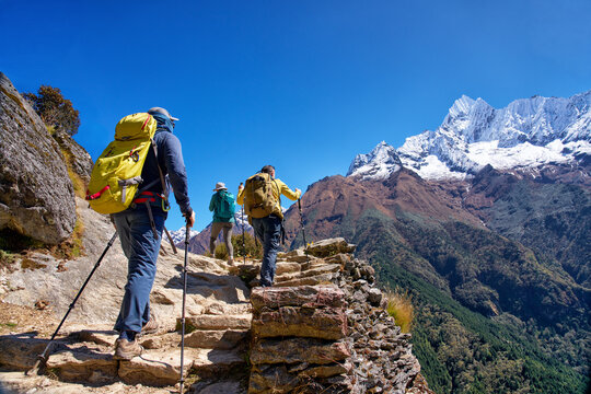 Group of hikers with backpacks and poles climbing ancient stone steps on trail to Everest Base Camp in Nepal, with snow-capped mountain peak.