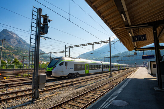 Modern BLS regional train arriving at Brig railway station platform under clear blue sky with Valais alpine mountain backdrop in Switzerland