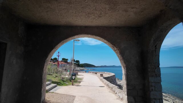Coastal pathway curving past a traditional stone building with an archway and red tiled roof. Surrounded by green Mediterranean pine trees and shrubs under a clear blue sky of Losinj Island in Croatia