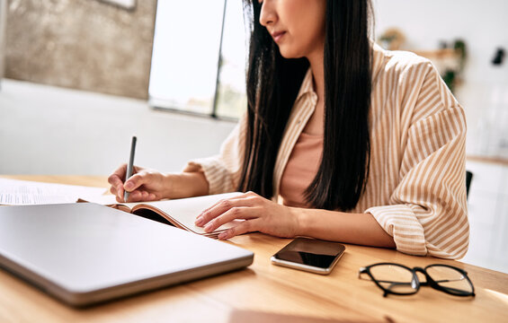 Young woman writing in a notebook at a desk with laptop, phone, and glasses, working on study or office tasks in a bright home setting.