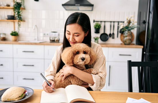 Woman hugging poodle while writing in kitchen.