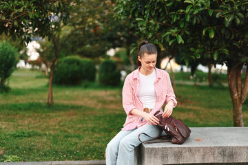 Obraz premium Woman lifestyle in a park sits on a bench wearing pink plaid shirt and jeans, adjusting a brown bag, calm outdoor moment, casual candid style