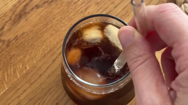 Hand stirring cold iced coffee with a glass straw to espresso in a tall cup, summer drink preparation