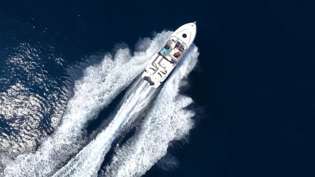 Aerial overhead tracking of a motorboat cruising with speed over the blue ocean and creating a trail of surf and waves