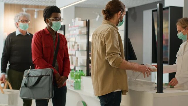 Multiethnic men wearing protective medical masks buying pills in pharmacy with Caucasian woman pharmacist seller female helping males buyers customers buy remedy medicine pandemic coronavirus outbreak