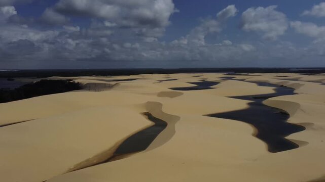 Sweeping drone shot over rolling golden sand dunes interspersed with dark freshwater lagoons under a cloudy sky in Lencois Maranhenses, Brazil.