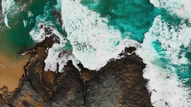 An abstract top-down aerial perspective capturing vibrant turquoise ocean waves crashing dramatically against dark, jagged basalt cliffs.