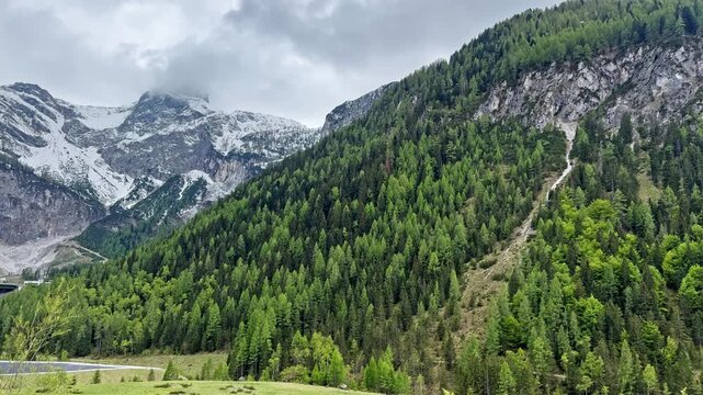 Alpine valley near Flachau with green forest, snowy Austrian peaks, rocky slopes, and cloudy mountain weather beside a highway route.