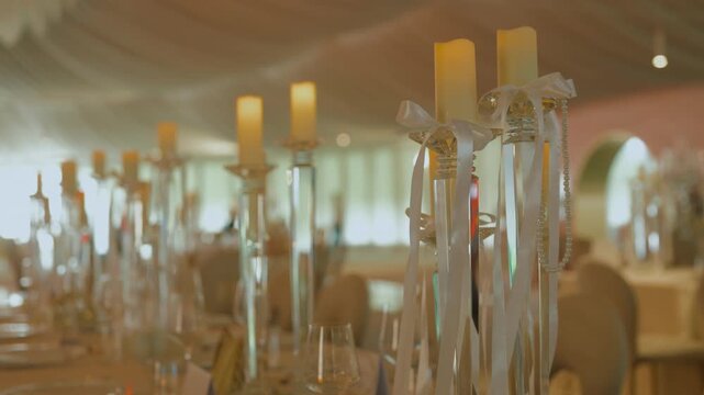 Close-up shot of a row of burning dinner candles placed on tall glass candlesticks decorated with white ribbons and pearl strings