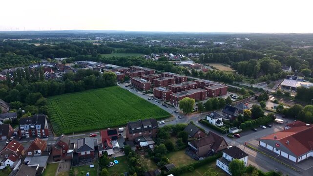Red brick apartment blocks border green farm field beside quiet German suburb at golden sunset, tree-lined streets and red-roof houses stretch across residential district. Aerial drone forward motion