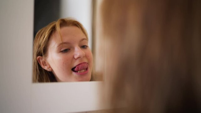 Happy teenage girl examining teeth and smiling at reflection in bathroom mirror. Healthy oral care routine, dental hygiene, self confidence, personal wellness and authentic everyday lifestyle.