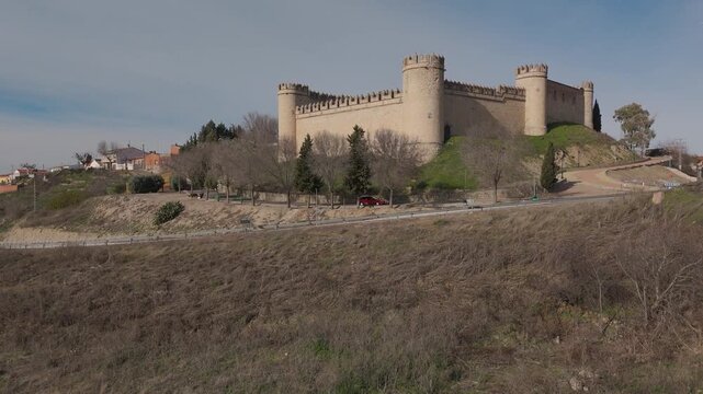 Ascending drone flight facing Maqueda Castle, showcasing its remarkably preserved medieval walls and pointed battlements. The fortress stands atop a mound beside the village on a sunny winter day.