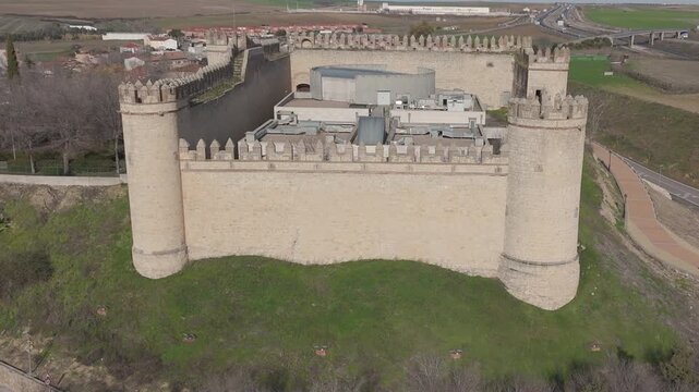 Front-facing drone flight approaching Maqueda Castle on a sunny winter morning, showcasing its imposing medieval walls, battlements, inner courtyard and the surrounding village and green fields
