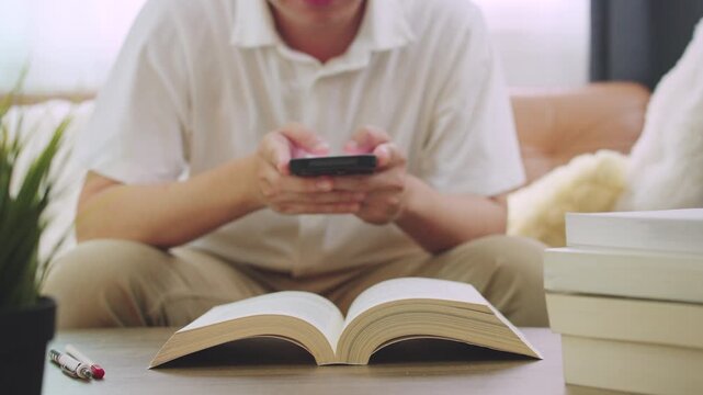 Student Man Taking a Break from Studying Siting on Couch Sofa Using Mobile Phone with Open book Foreground.