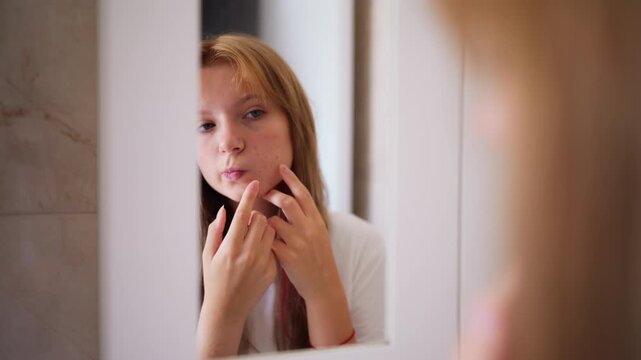 Worried young woman in her bathroom examining her face in the mirror, squeezing a pimple. Unhappy teenager suffering from acne and skin problems during puberty