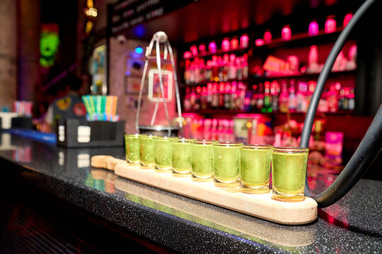 Row green shot glasses on wooden serving board next to glass cocktail shaker on dark bar counter blurred vibrant neon pink red lights creating lively nightclub party atmosphere for social celebration