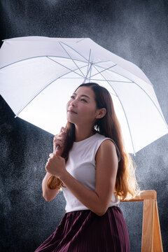 Beautiful Asian woman sitting holding white umbrella under rain on dark background. Happy female looking up enjoying water drops. Weather protection and insurance concept with copy space.