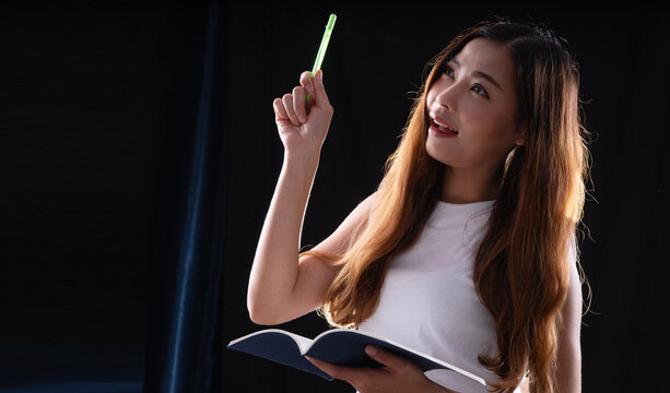 Smiling young Asian woman holding notebook and pointing pen up on dark background with copy space. Happy female student or businesswoman getting inspiration or creative idea.