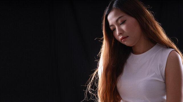 Sad lonely young Asian woman in white top looking down with depressed expression on dark black background with copy space. Mental health, stress, sorrow and depression concept.