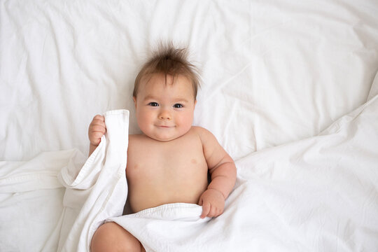 A cute nine-month-old baby boy is lying on a bed on white sheet, smiling and playing with a blanket. A funny chubby baby. A real photo, real baby, live emotions. Minimalist clean background, copy spac