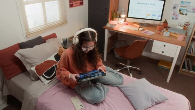 Caucasian teenager wearing headphones sitting on bed and using tablet for online studying. Clip is ideal for e-learning platforms, educational apps, and digital student lifestyle campaigns