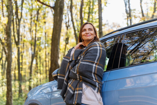 Cheerful mature woman wrapped in plaid blanket taking a break near vehicle, enjoying autumn sunshine. Healthy aging concept