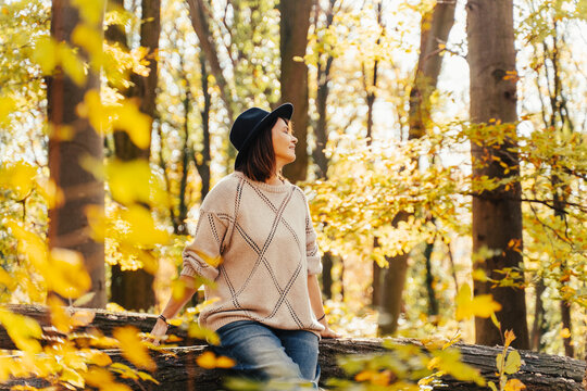 Beautiful thoughtful mature woman in fashion hat and cozy clothing looking away, sitting on nature during autumn weekend.