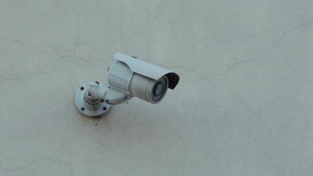 Wall mounted security camera watching over plain stucco surface with weathered metal housing and urban surveillance feel