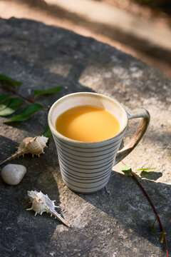 Overhead-style outdoor composition with a striped ceramic cup of orange juice, fresh peaches in a matching bowl, seashells, pebble and green leaves on stone.